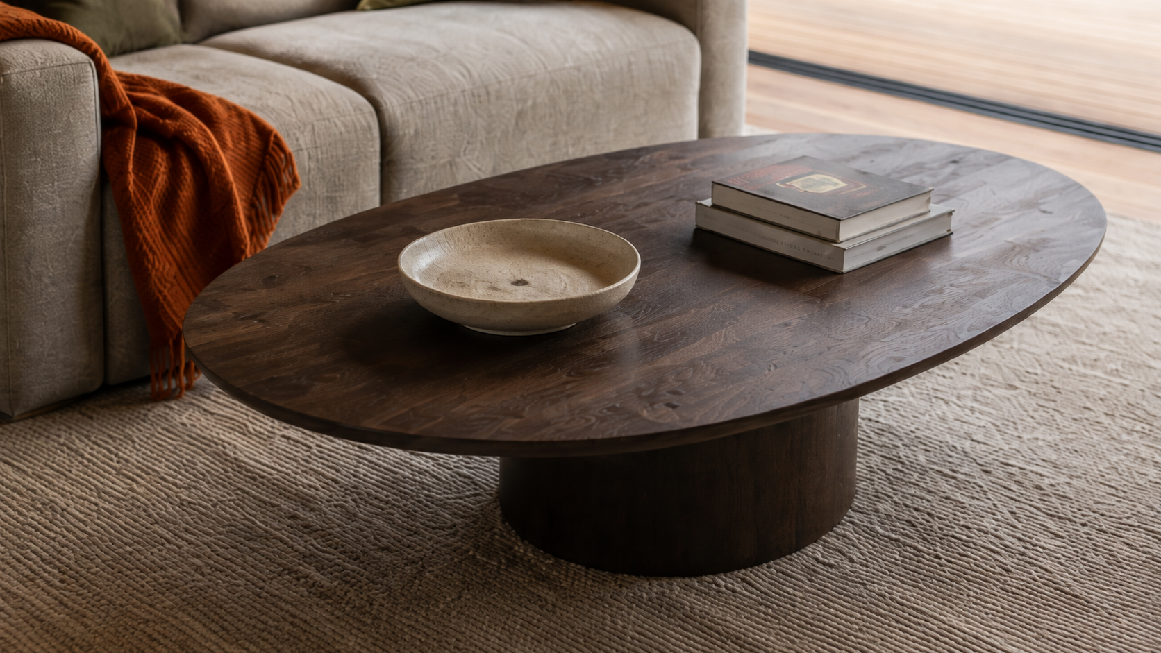 AI-generated Concept close living-room detail of the oval timber table and broad pedestal, shallow cream bowl, two closed books, grey sofa, rust throw and woven rug.